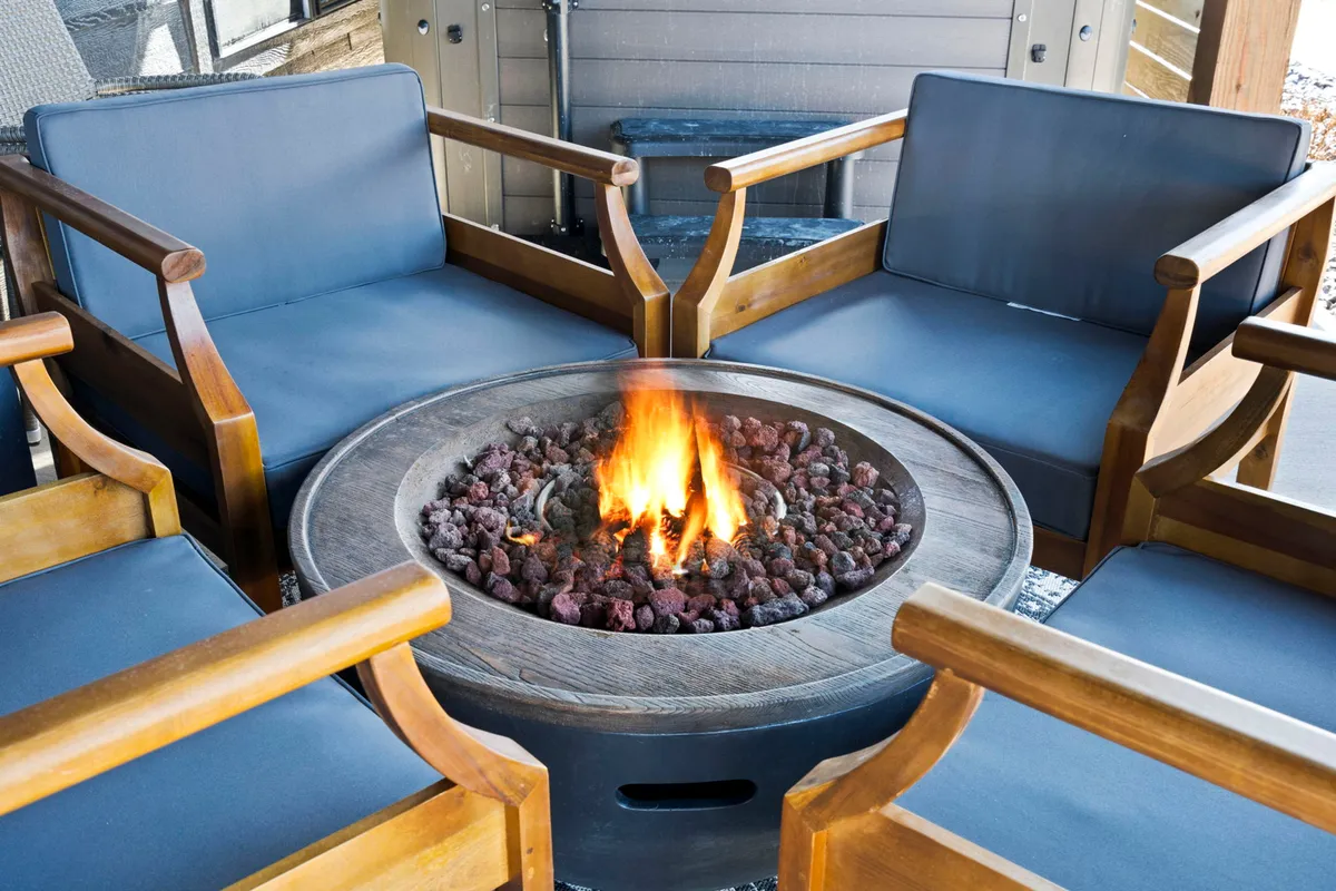 An exterior evening scene featuring a cozy fire pit surrounded by wooden chairs and rustic rock decorations. The seating area invites guests to gather, relax, and enjoy the outdoors under the stars, evoking feelings of warmth and togetherness.