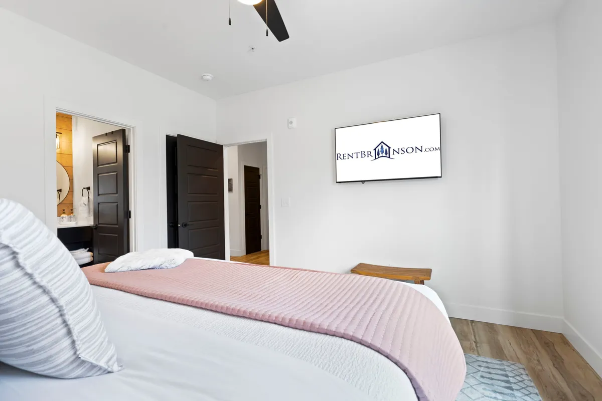 A tranquil bedroom with a comfortable bed dressed in a pink throw, a television mounted on the wall, and a doorway leading into a connected bathroom. The ambiance is comfortable and private, with the TV highlighting personal leisure time.
