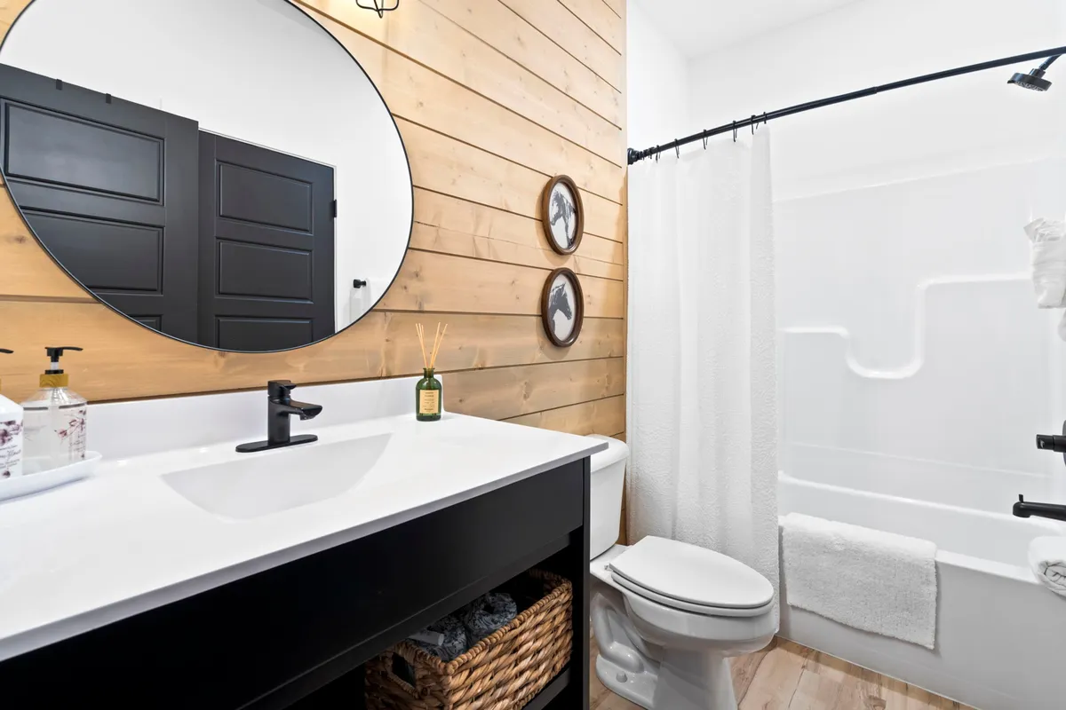 A contemporary bathroom with a round mirror above a black sink and vanity, a wooden wall, and a shower curtain. The scene is calm and well-lit, ideal for refreshing morning or evening routines.