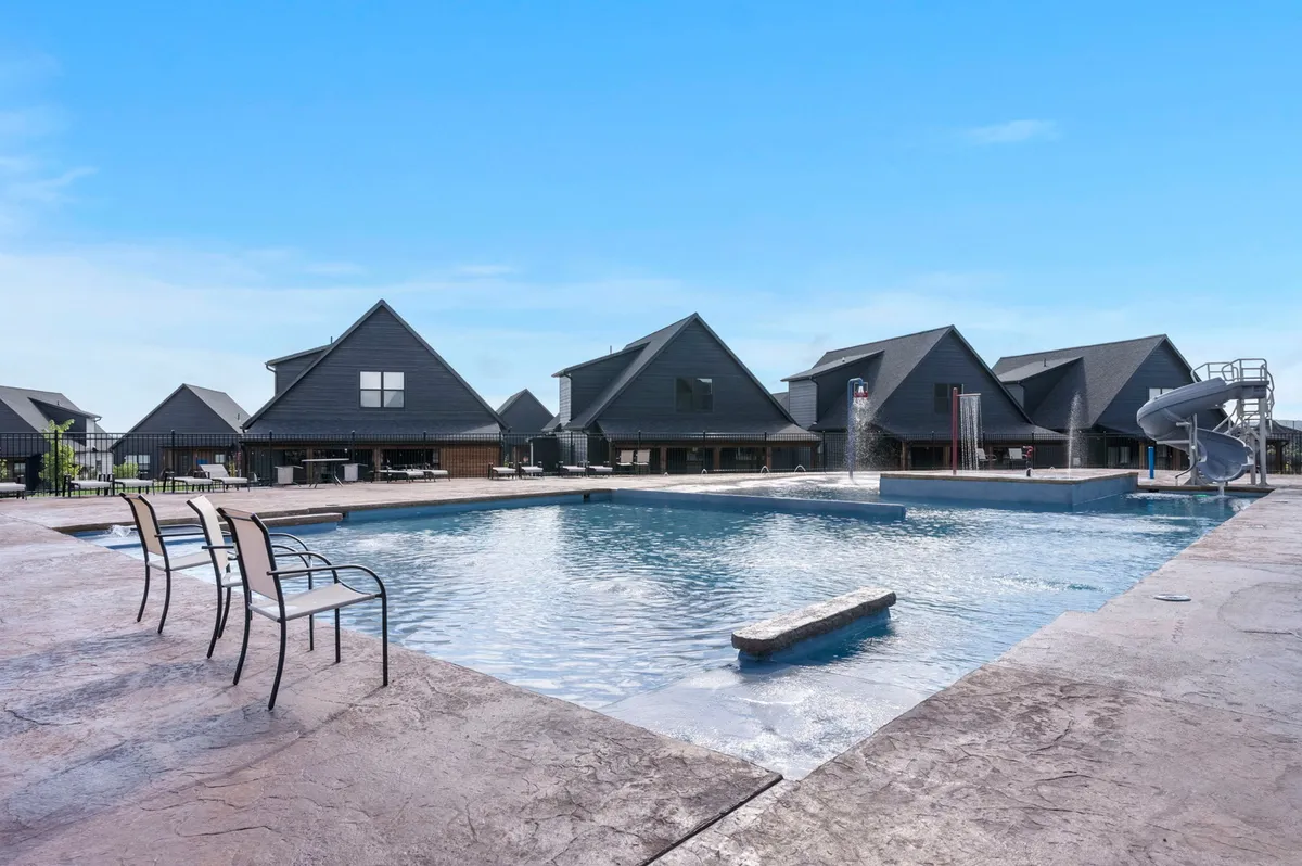 A wide exterior shot of the resort-style swimming pool, complete with a fun water slide, empty lounge chairs ready for guests, and neighboring houses in the background. The scene is bathed in warm, late-afternoon sunlight, creating an inviting atmosphere for families to relax and play.