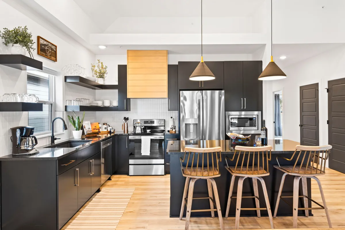 The image showcases a stylish, open-concept kitchen with a central island featuring comfortable bar stools, sleek black cabinetry, stainless steel appliances, and open shelving with neatly arranged glassware. The space is bathed in soft natural light, creating an inviting atmosphere ideal for social gatherings, family breakfasts, or relaxed evenings in.