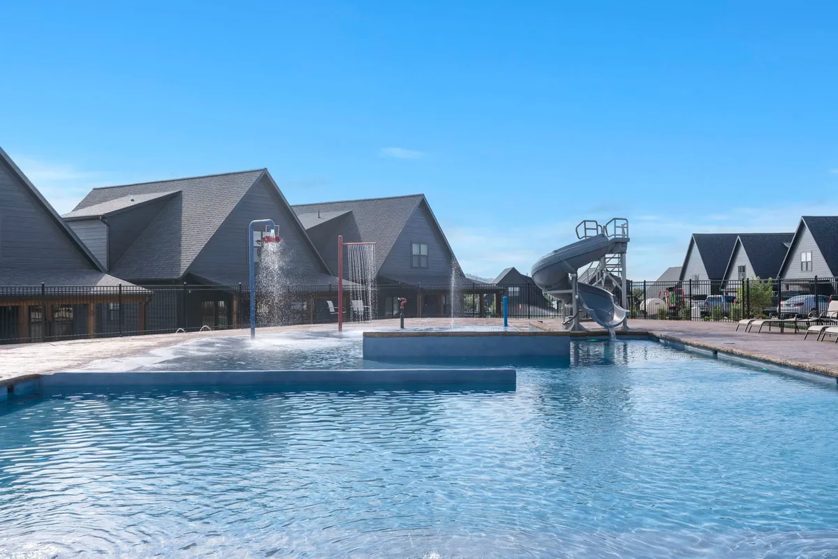 A vibrant exterior shot of the shared resort-style swimming pool, complete with a water slide and splash pad. Surrounding buildings and empty lounge chairs hint at easy access and exclusive community perks, while the overall mood is playful and inviting for families and kids.