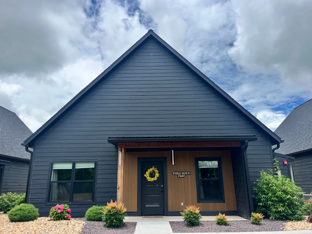 An inviting exterior scene with a front door adorned with a yellow wreath, wooden accents, and landscaped flower beds. The porch and garden setting hints at relaxing and welcoming outdoor moments.