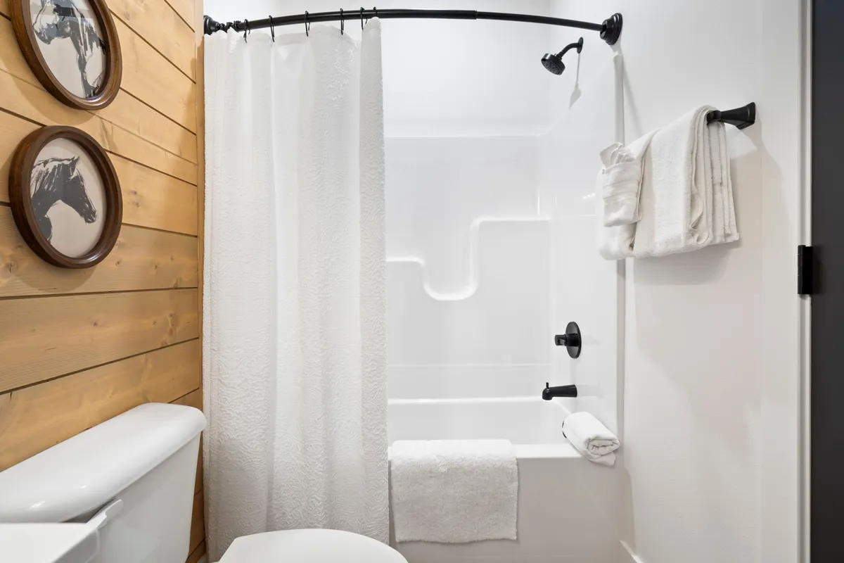 A modern ensuite bathroom showcasing wooden wall panels, a combined bathtub and shower area, towels on a rack, and unique horse artwork above the tub. The mood is calm and inviting, ideal for relaxation after a day of lake activities.