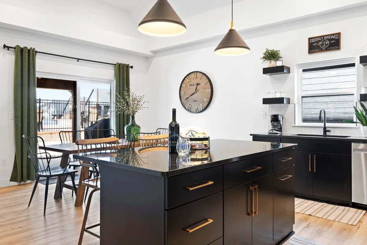 A warm, welcoming kitchen and dining area featuring a large island, elegant pendant lighting, a wall clock, and high-backed dining chairs. The space is set up for hosting, cooking, and casual family meals.