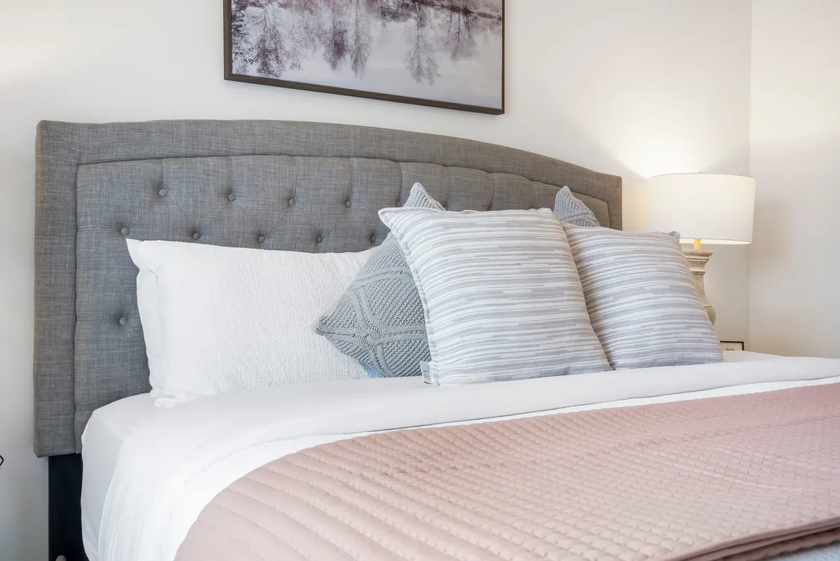 A master bedroom featuring a plush king bed with a grey upholstered headboard, decorative pillows, a soft pink bedspread, a nightstand with a lamp, and framed wall art. The overall ambiance is serene and cozy, perfect for restful nights or calm mornings.