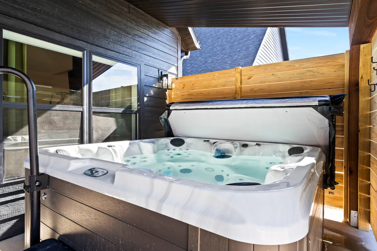 A private hot tub set outdoors, partially enclosed by wooden fencing for privacy, with the home's glass windows reflecting sunset colors. The ambiance is inviting and serene, ideal for unwinding while soaking in views of the Ozark cliffs or stargazing at night.